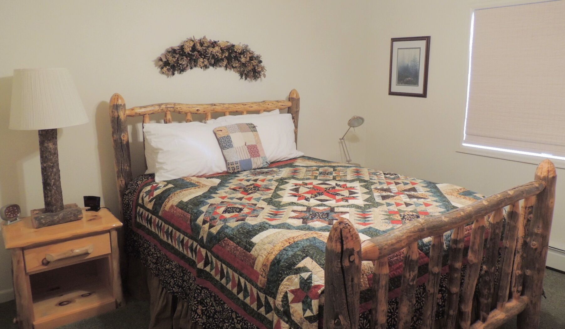 A bedroom with a wooden bed frame, a quilt, a nightstand, a lamp, a wall decoration, and a framed picture.