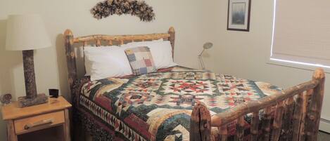 A bedroom with a wooden bed frame, a quilt, a nightstand, a lamp, a wall decoration, and a framed picture.
