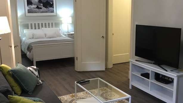 A living room with a sofa, a coffee table, a TV on a stand, a bed with a headboard, and a door leading to another room.