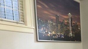 A dining table with white chairs, two bowls, and wine glasses set for two. A cityscape painting hangs above the table.