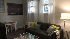 A living room with a dark sofa, a glass coffee table, a dining table with chairs, and a cityscape painting on the wall.