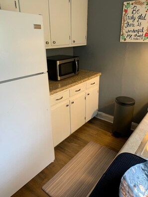 Compact kitchen area with a refrigerator, microwave, and trash bin, featuring a rug and a motivational quote on the wall.