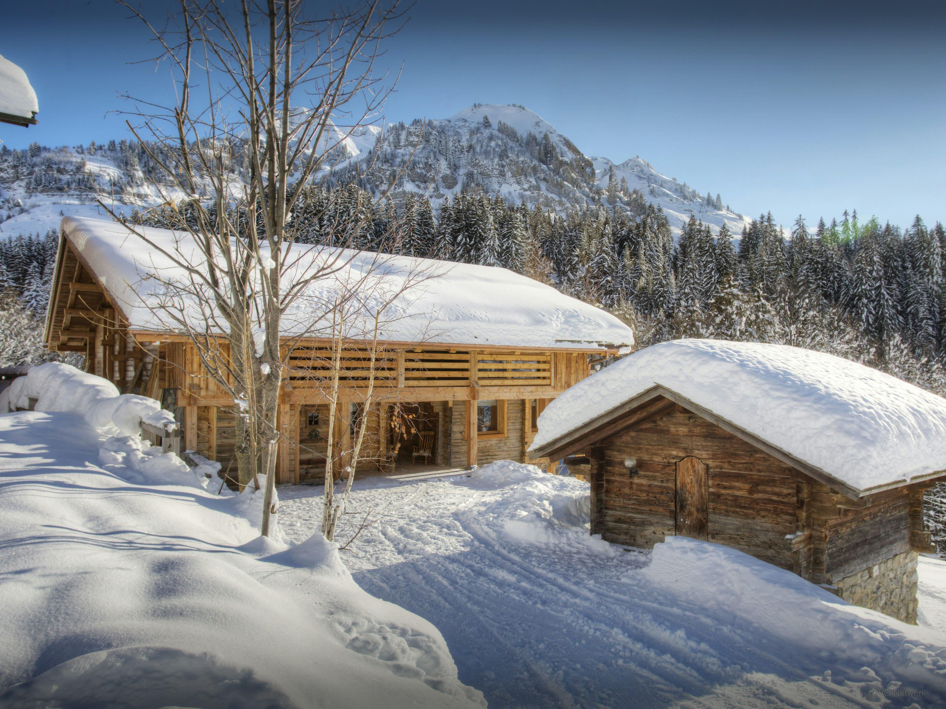 A wooden cabin with a snow-covered roof, surrounded by a snowy landscape and a mountain in the background.