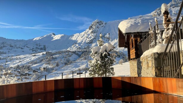 A wooden bridge over a reflective water surface, leading to a building with a stone facade and snow-covered surroundings. - Chalet Argali - luxury ski-in ski-out base for 14 - OVO Network (Le Grand-Bornand)