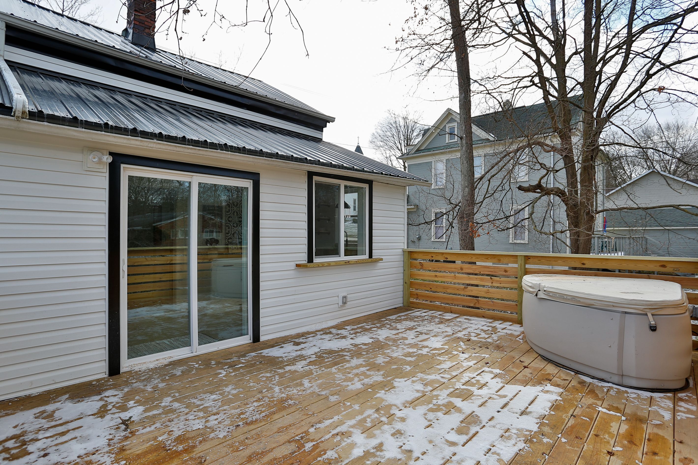 A covered deck with a hot tub, wooden flooring, and a sliding glass door leading to a room with a metal roof.