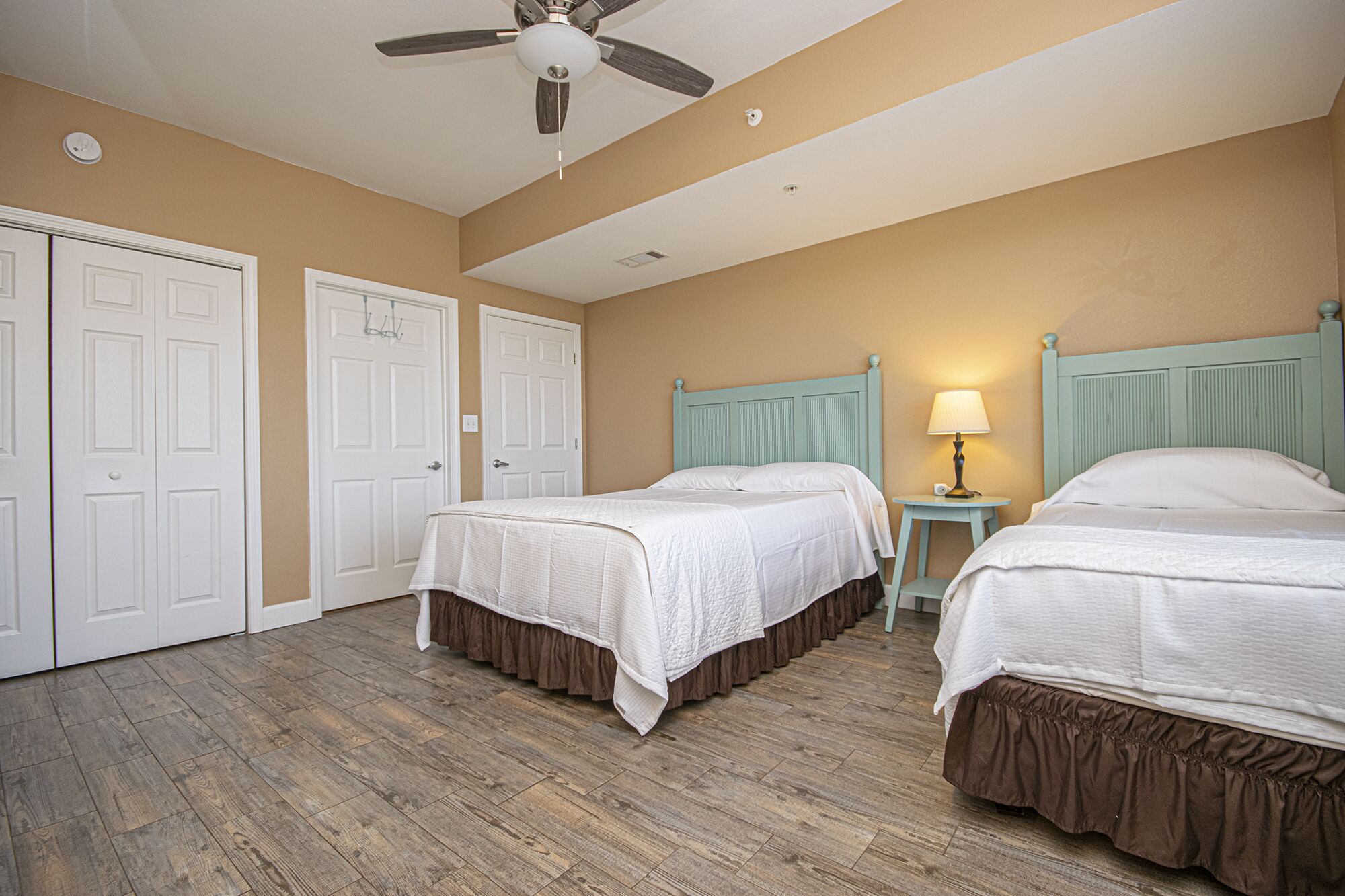 A hotel room with two beds, a ceiling fan, a nightstand with a lamp, and a door.