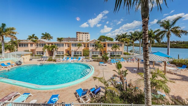 A resort with a swimming pool, lounge chairs, palm trees, and a building with multiple floors.