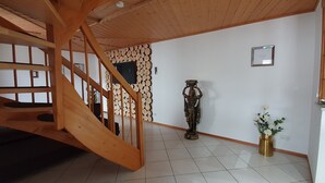 Un escalier en bois mène à une pièce dont le sol est recouvert de carreaux, où l’on trouve un téléviseur fixé au mur et une statue décorative.