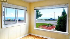 A room with two large windows, wooden flooring, and a view of the sea and a parking lot. - BAYVIEW AND SUNRISE! 1 bedroom close to ski hills and Bruce County (MEAFORD)