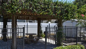 Un espacio de estar al aire libre con techo, con una mesa de picnic y bancos, rodeado por una valla de metal y follaje.