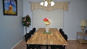 Un comedor con una mesa de madera, sillas negras, un jarrón con flores, una pared con un cartel y una ventana con persianas.