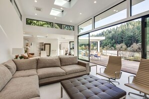 Una sala de estar moderna con un sofá modular, un reposapiés de cuero y un amplio ventanal con vistas a un área boscosa.