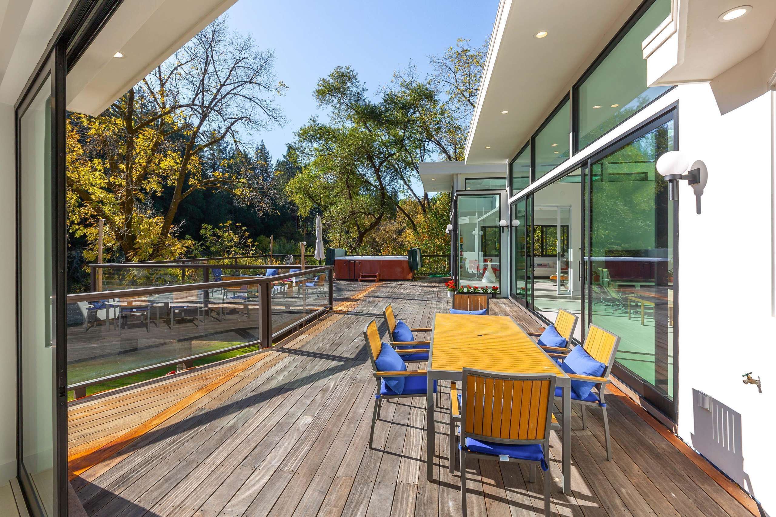 Una terraza moderna al aire libre con mesa y sillas de madera, barandillas de vidrio y vistas a árboles y follaje.