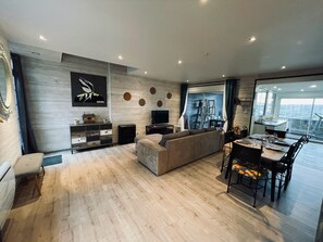 A modern living room with a large sofa, a dining area with a table and chairs, a TV mounted on the wall, and a view of the outside through a sliding door. - La BOUGERIE de Chaligny (Chaligny)