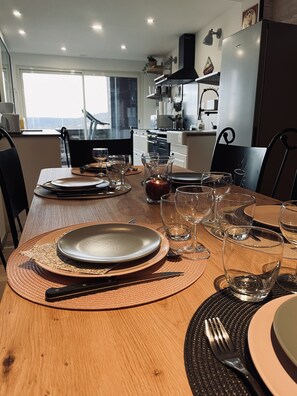 A dining table set with plates, cutlery, and glasses, with a candle and a view of the kitchen and living area in the background. - La BOUGERIE de Chaligny (Chaligny)