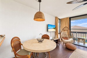 A modern living room with a round table, wicker chairs, a hanging light fixture, a television, and a large window with curtains.