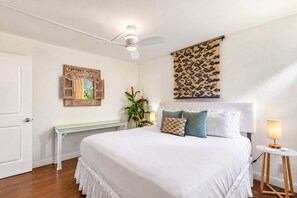 A hotel room with a large bed, a ceiling fan, a wall-mounted tapestry, a wooden bench, and a bedside table with a lamp.