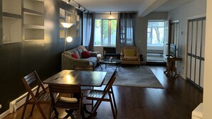 A modern living room with a sofa, armchair, dining table, and chairs. There is a floor lamp, a small table with items, and a view of the outside through a window.