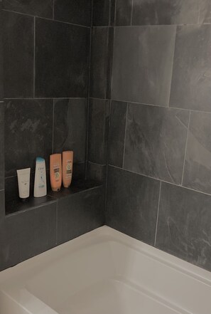 Une salle de bain avec des carreaux de couleur foncée, une baignoire blanche et trois produits de toilette disposés sur une étagère.
