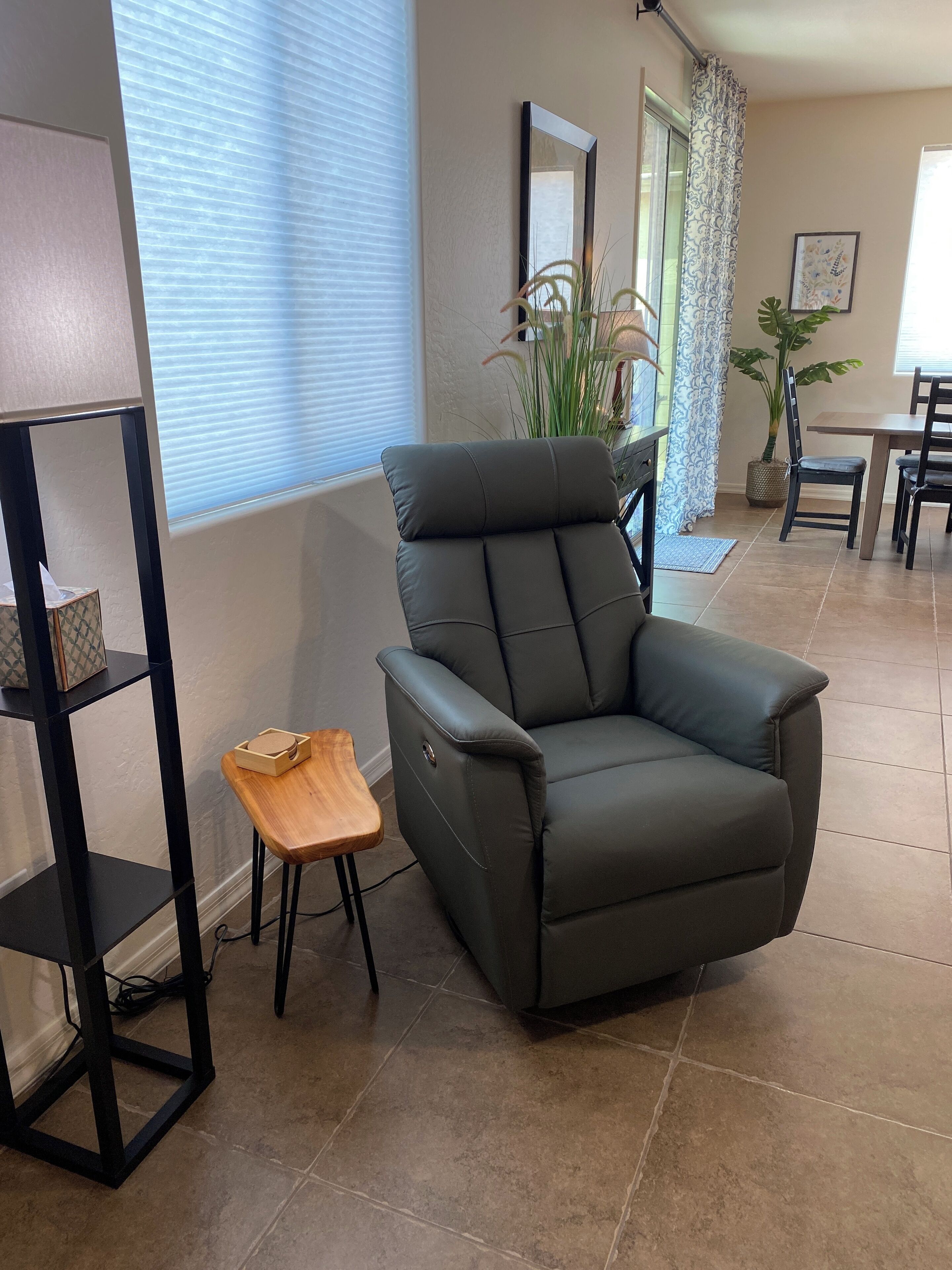 A living room with a grey armchair, a wooden side table, a black shelving unit, a dining table with chairs, and a potted plant.