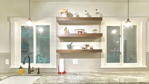 A kitchen with white cabinets, a stainless steel dishwasher, wooden shelves with kitchen items, and pendant lights.