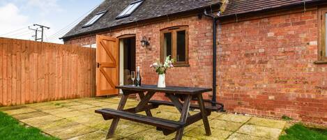 A wooden picnic table in a backyard with a brick house, a wooden fence, and a clear sky.