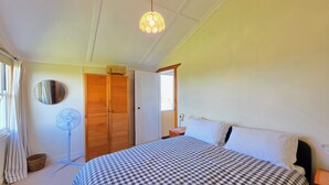 Une chambre avec une couvre-lit à carreaux, un ventilateur sur pied, un miroir, une armoire en bois et un luminaire fixé au plafond.
