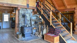 A cozy cabin interior with a stone fireplace, a wooden staircase, and a log storage area. - Green Acres Ranch Family Style (Thompson Falls)