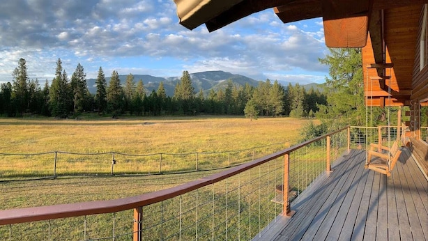 A wooden deck with a metal railing overlooking a grassy field and trees, with mountains in the background. - Green Acres Ranch Family Style (Thompson Falls)
