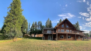 A large wooden house with a balcony, surrounded by a grassy field and tall trees. - Green Acres Ranch Family Style (Thompson Falls)
