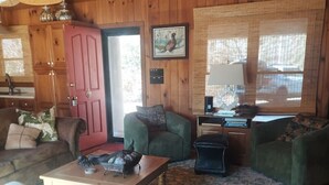 L’intérieur d’un chalet accueillant, avec des murs en bois, une table à manger, deux fauteuils verts, un canapé et une petite table d’appoint.