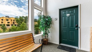 A bench, a green door, and a potted plant in a hallway with a view of buildings outside.