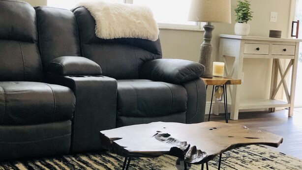 A modern living room with a black leather sofa, a white fluffy throw, a wooden coffee table, a lamp, and a small plant on a white console table. - Prime Location in The Villages – Golf Cart Included and 2 King Sized Beds! (The Villages)