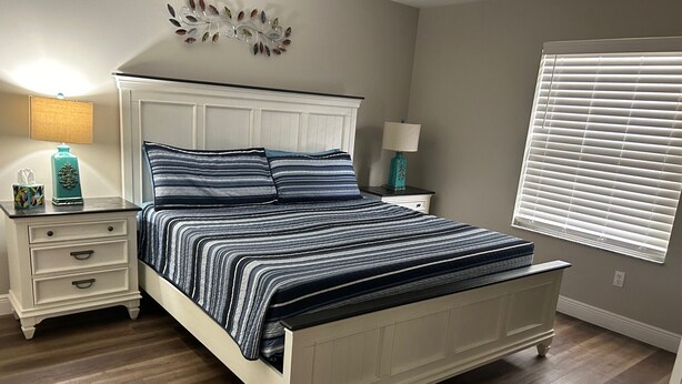 A bedroom with a bed, nightstands, a wall decoration, and a window with blinds. - Prime Location in The Villages – Golf Cart Included and 2 King Sized Beds! (The Villages)