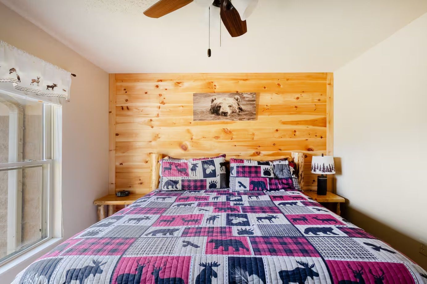 A bedroom with a wooden headboard, a quilted bedspread, a nightstand with a lamp, and a window with curtains.