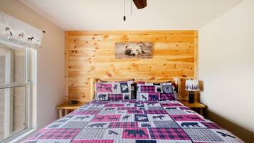 A bedroom with a wooden headboard, a quilted bedspread, a nightstand with a lamp, and a window with curtains.