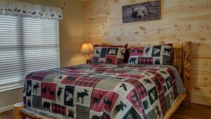 Un dormitorio acogedor con cabecera de madera, colcha acolchada con estampados de animales, lámpara de pared y ventana con persianas.