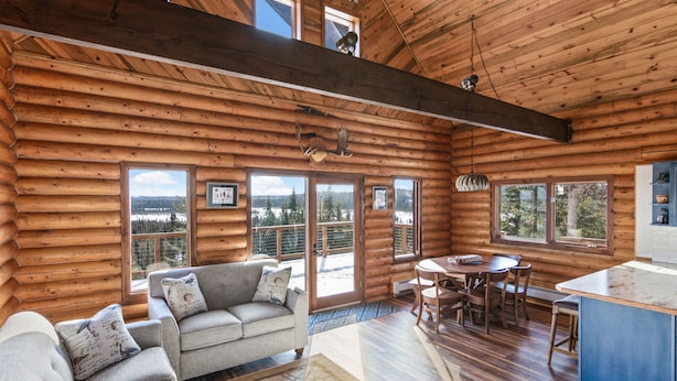 A log cabin interior with a dining area, sofa, and large windows offering views of a snowy landscape.