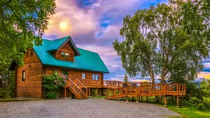 A log cabin with a green roof, a wooden deck, and a gravel driveway.