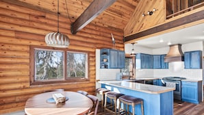 A wooden cabin interior with a round dining table, bar stools, a kitchen with blue cabinets, and a large window overlooking trees.