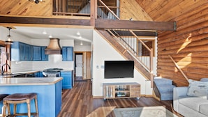 A spacious living area with a large TV, wooden staircase, and a kitchen with blue cabinets.