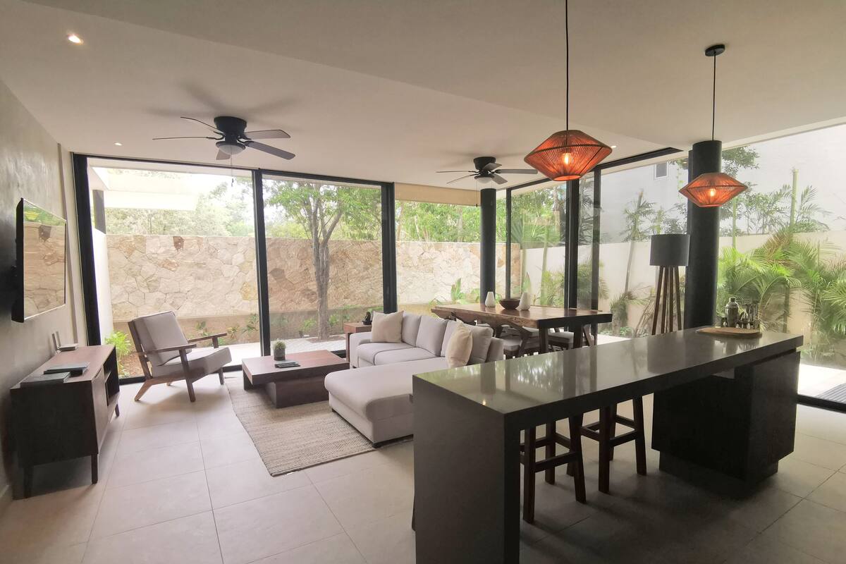 Una sala de estar moderna con una isla grande, sofás y un comedor. Hay ventiladores de techo y lámparas colgantes.