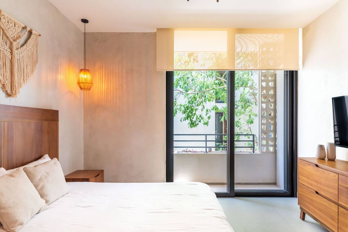 Un dormitorio moderno con una cama, una mesita de noche de madera, un adorno de macramé en la pared, una lámpara colgante, un amplio ventanal con vistas a la vegetación y un televisor sobre un mueble de madera.