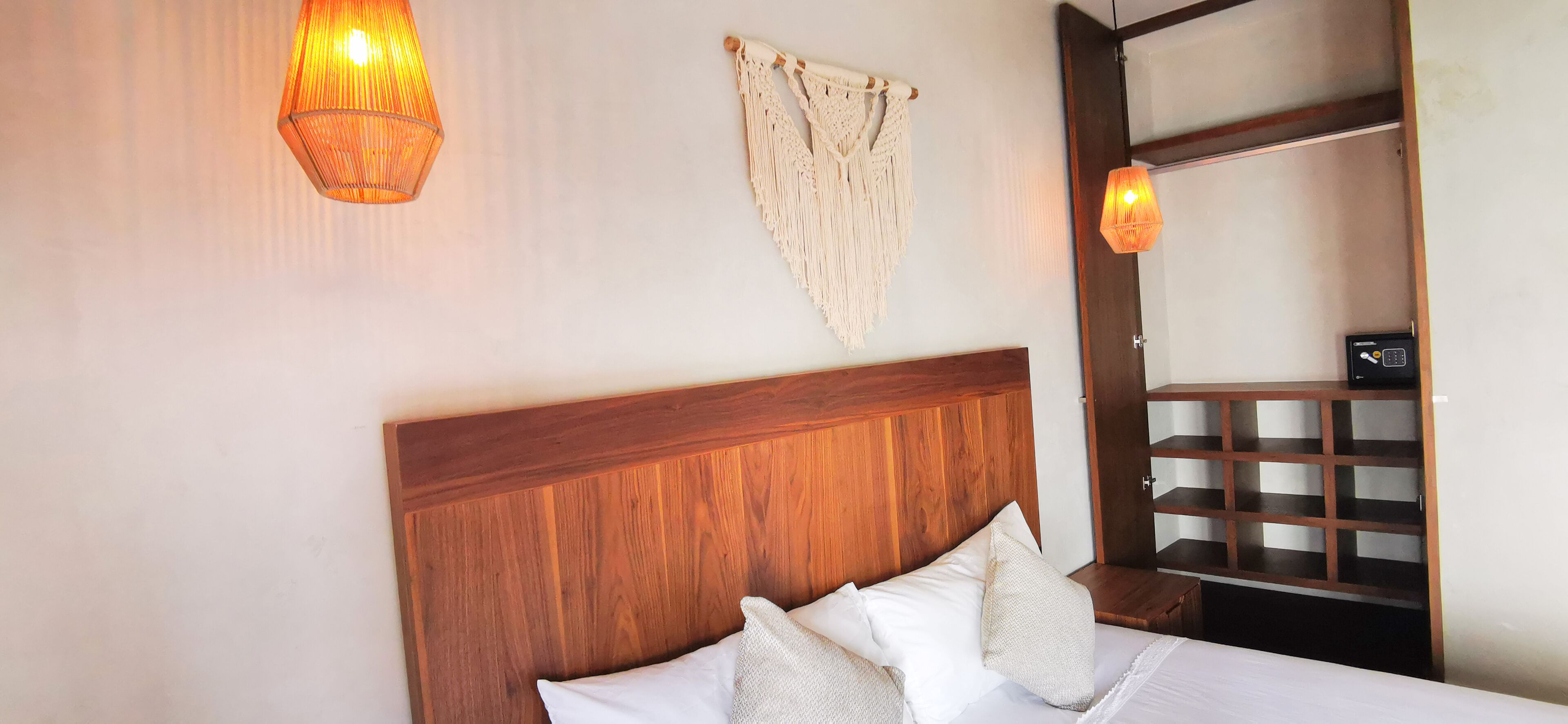 Una cama bien hecha con cabecera de madera, dos luces fijadas en la pared y un tapiz de macramé.