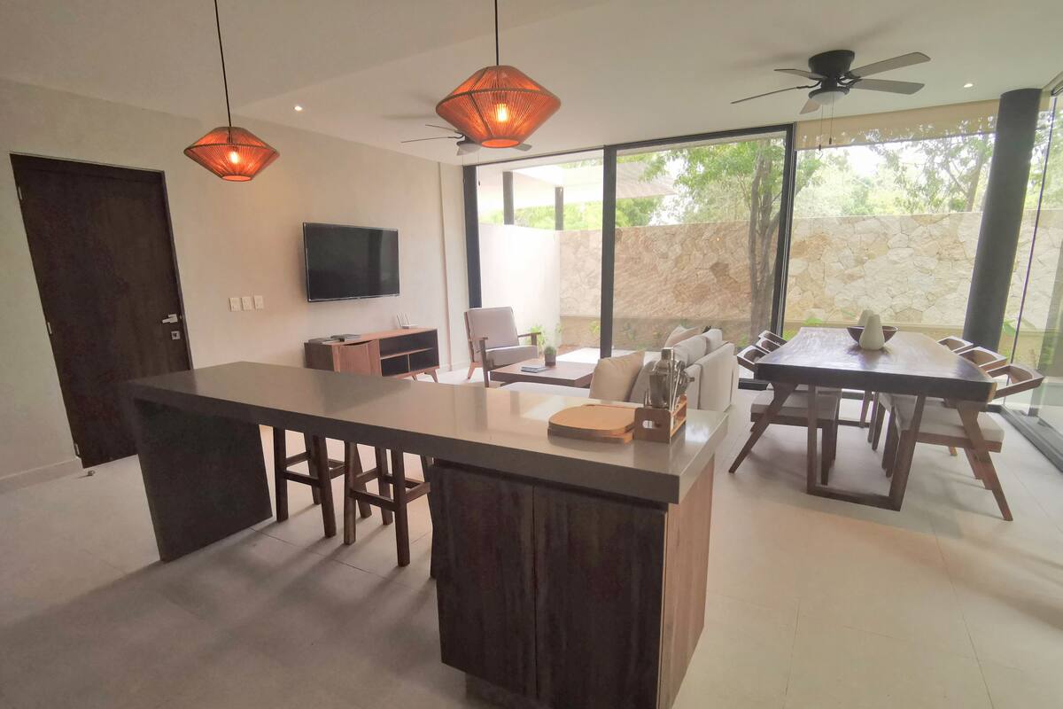 Una cocina moderna con una isla central, un comedor con mesa y sillas, un televisor de pantalla plana montado en la pared y ventanas amplias con vistas al exterior.