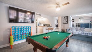 Une table de billard avec des billes, une queue de billard et un flipper coloré dans un salon équipé d’un canapé et d’un téléviseur fixé au mur.