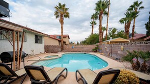 Un jardin avec une piscine, des chaises longues, des palmiers et un barbecue.