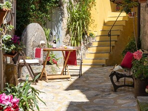 A stone-paved courtyard with a wooden table set for a meal, surrounded by potted plants and a red chair.