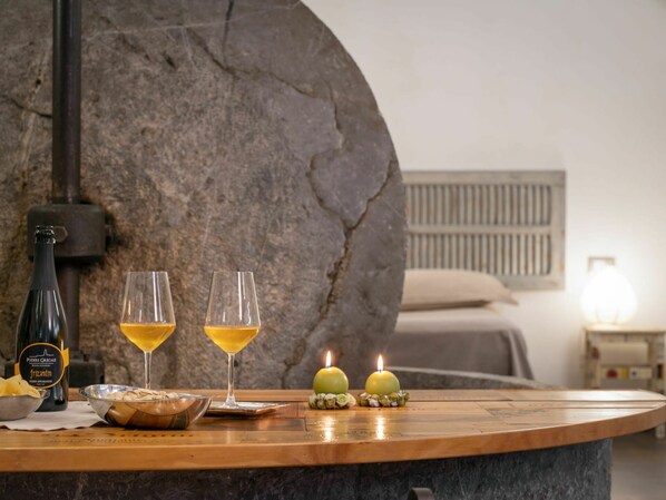 A wooden table with two glasses of wine, a bottle, and candles in a room with stone walls and a bed in the background.