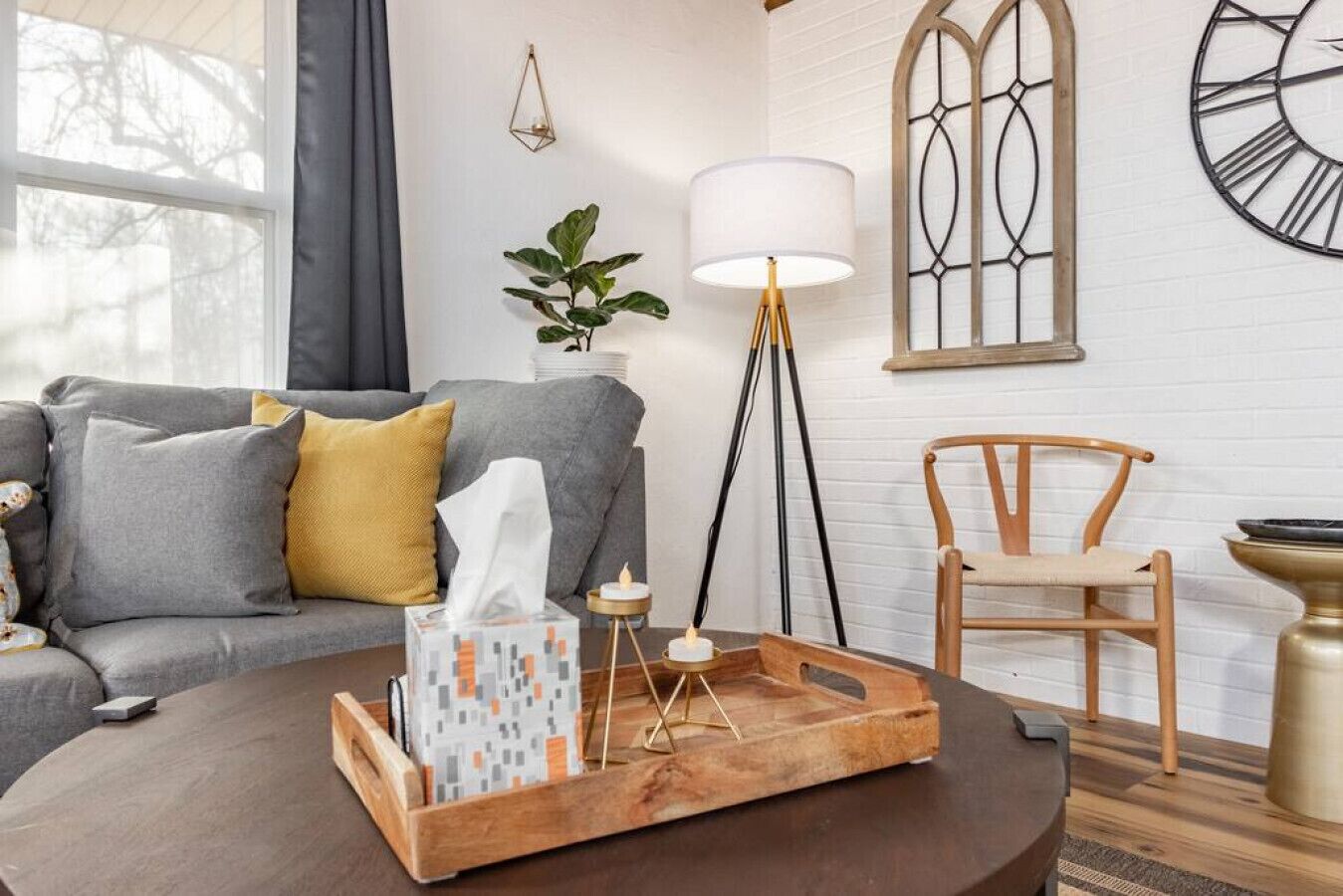 Een woonkamer met een grijze bank, een houten bijzettafeltje, een vloerlamp en een decoratieve wandklok.